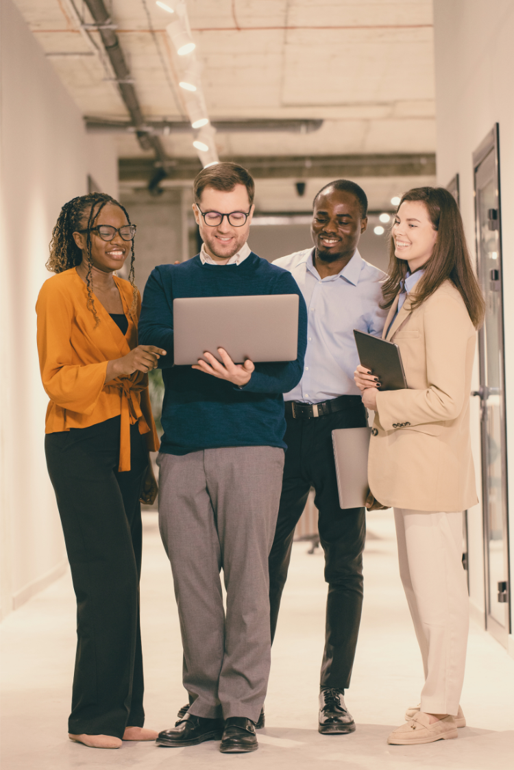 Four professionals collaborate in a modern office hallway, reviewing information together on a laptop.